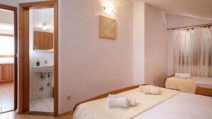 A hotel room with a wooden floor, a large bed with a white bedspread, two bedside tables with towels, a bathroom with a sink and mirror, and a painting on the wall. - Dolac Green Side Apartments (Jelsa)