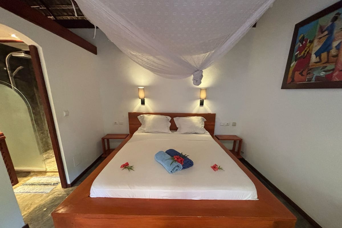 Una cama bien hecha con edredón blanco, dos almohadas y una toalla azul con flores. Hay dos luces empotradas en la pared y un cuadro colgado.