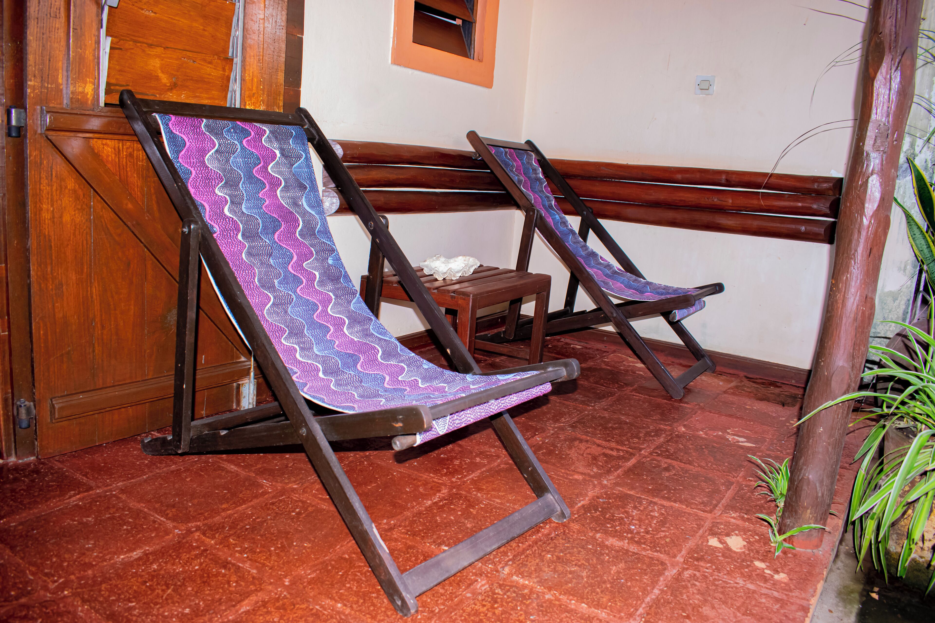 Two wooden lounge chairs with colorful fabric backs, positioned on a red-tiled floor, adjacent to a wooden door and a small window.