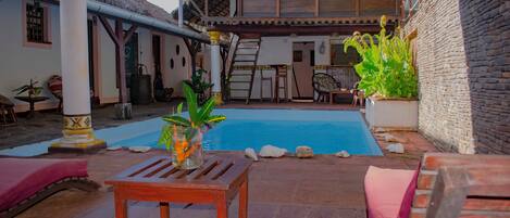 A pool area with a wooden table and lounge chairs, a staircase leading to an upper level, and a stone wall.