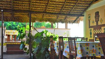 An outdoor dining area with wooden tables and chairs, covered by a thatched roof, and surrounded by lush greenery.