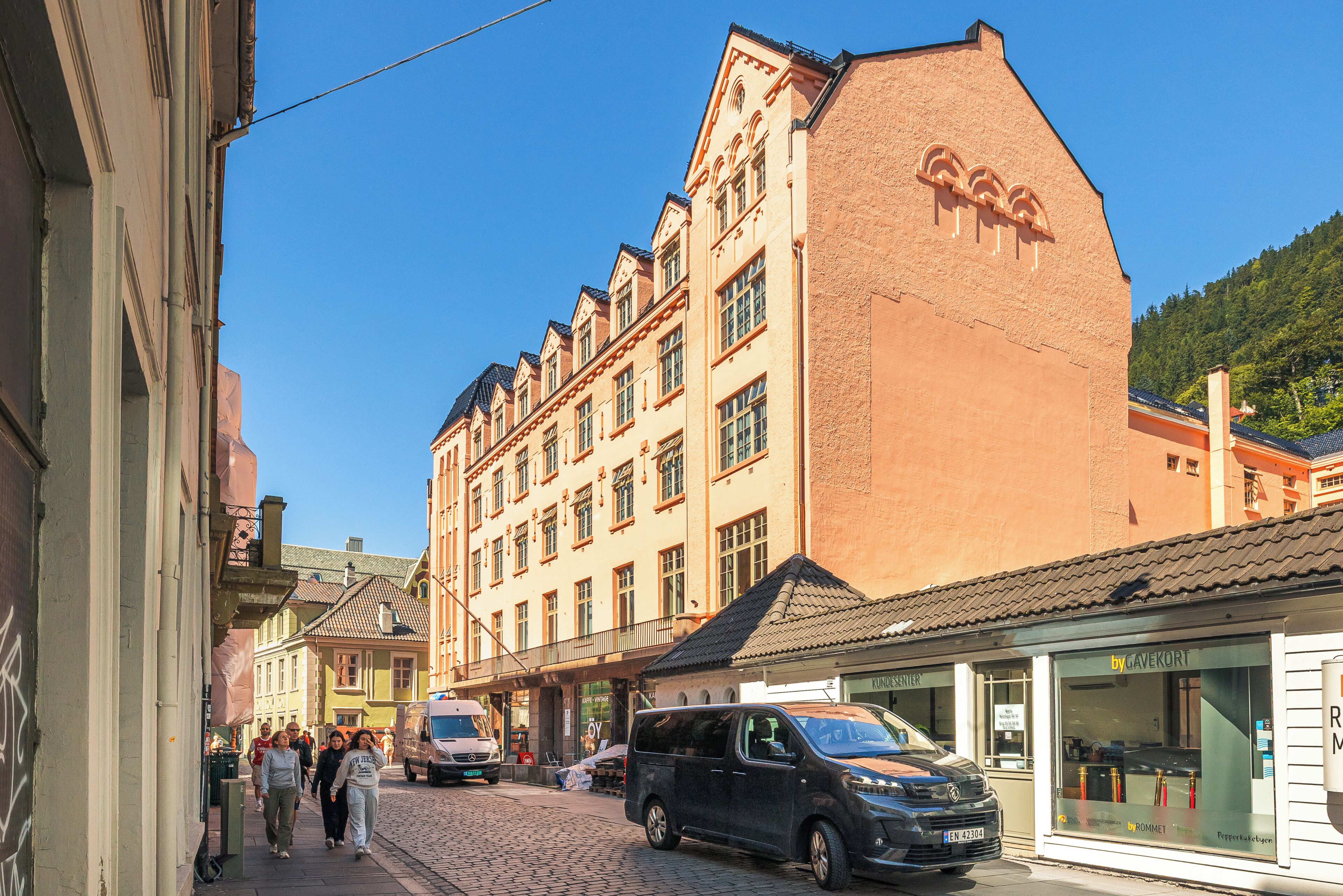 A cobblestone street lined with historic buildings, a black van parked on the side, and pedestrians walking.