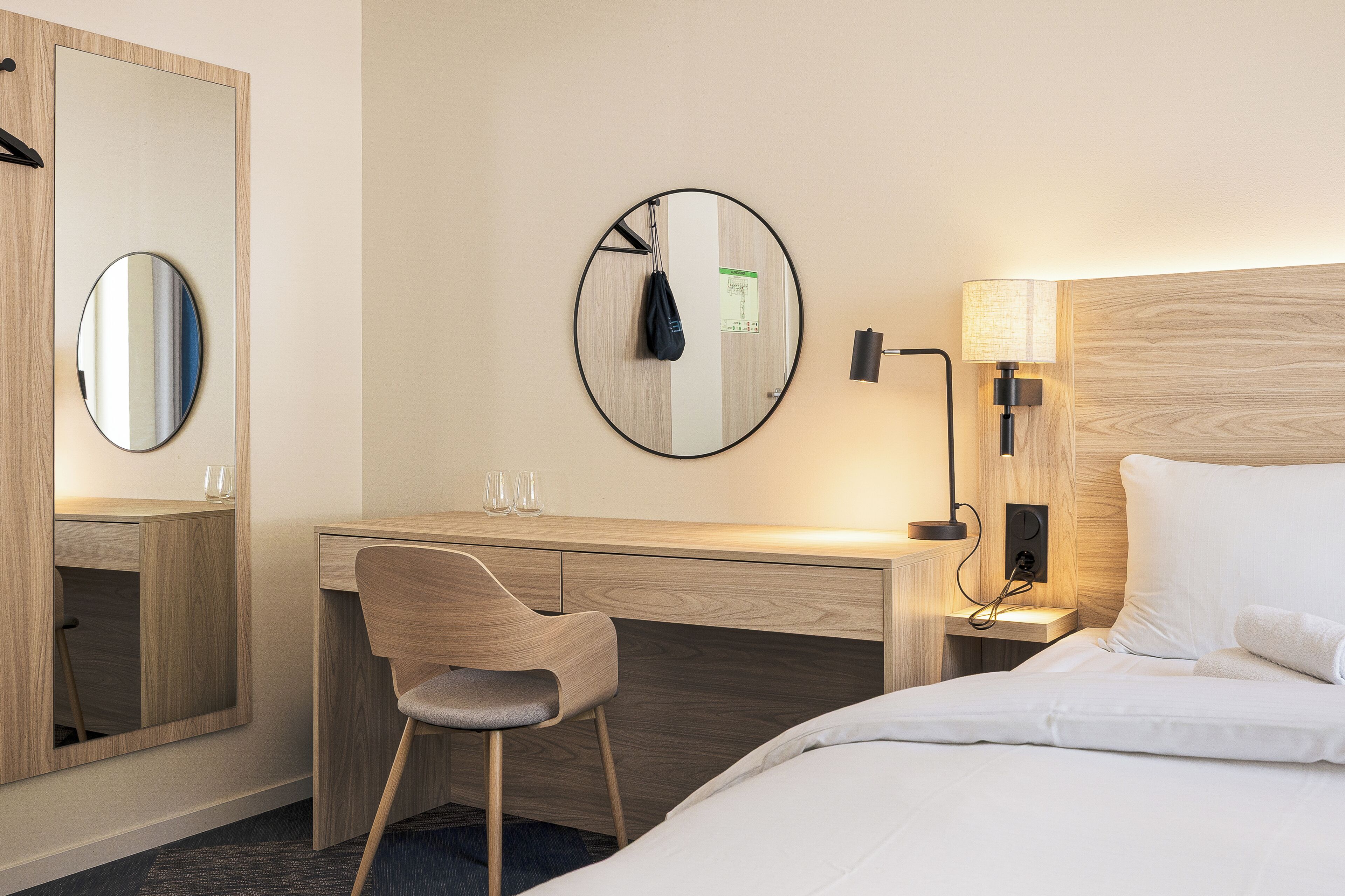 Hotelkamer met een bed, een bureau met stoel, een ronde spiegel en een wandlamp.