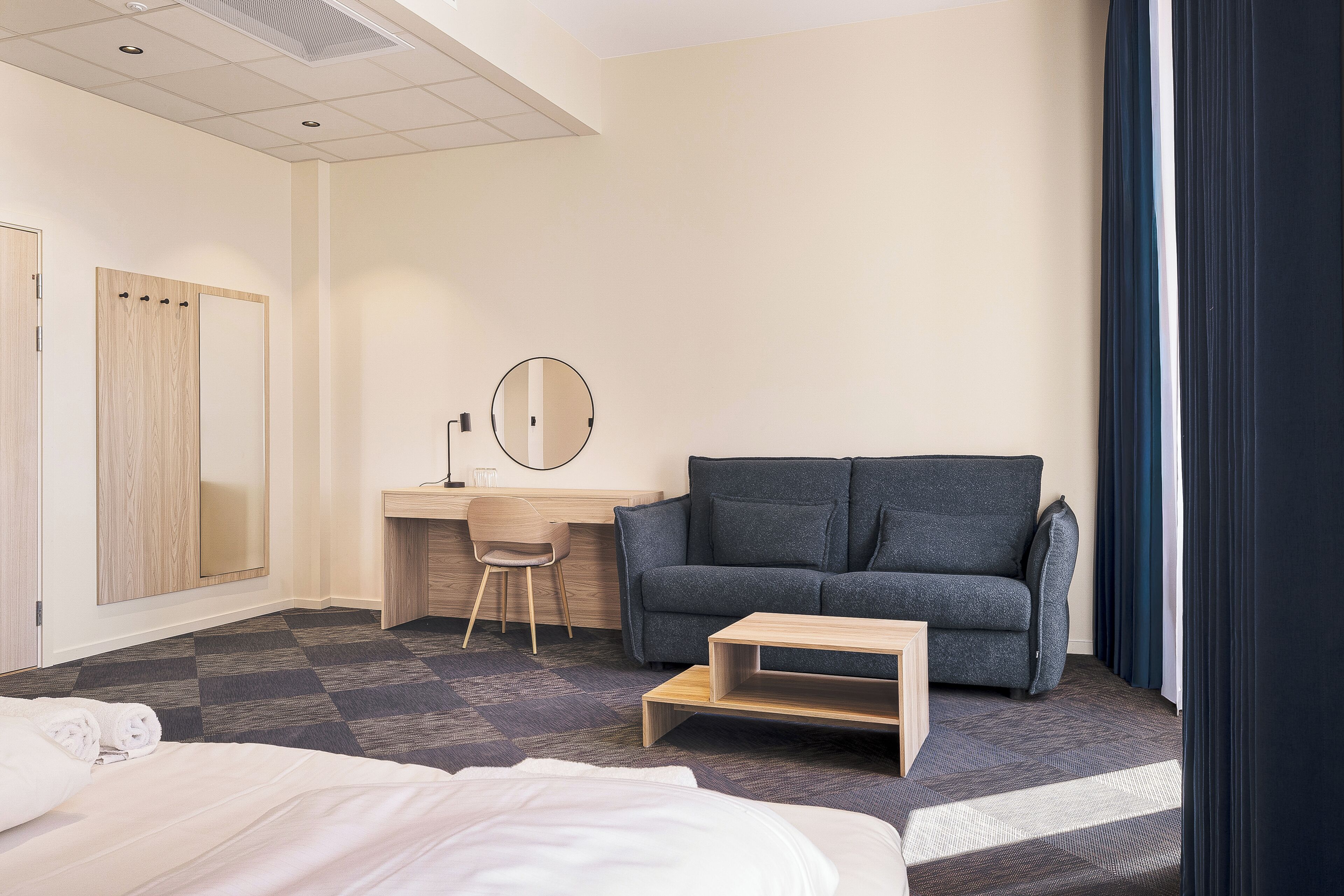 A hotel room with a bed, a desk with a chair, a sofa, a small table, and a mirror.