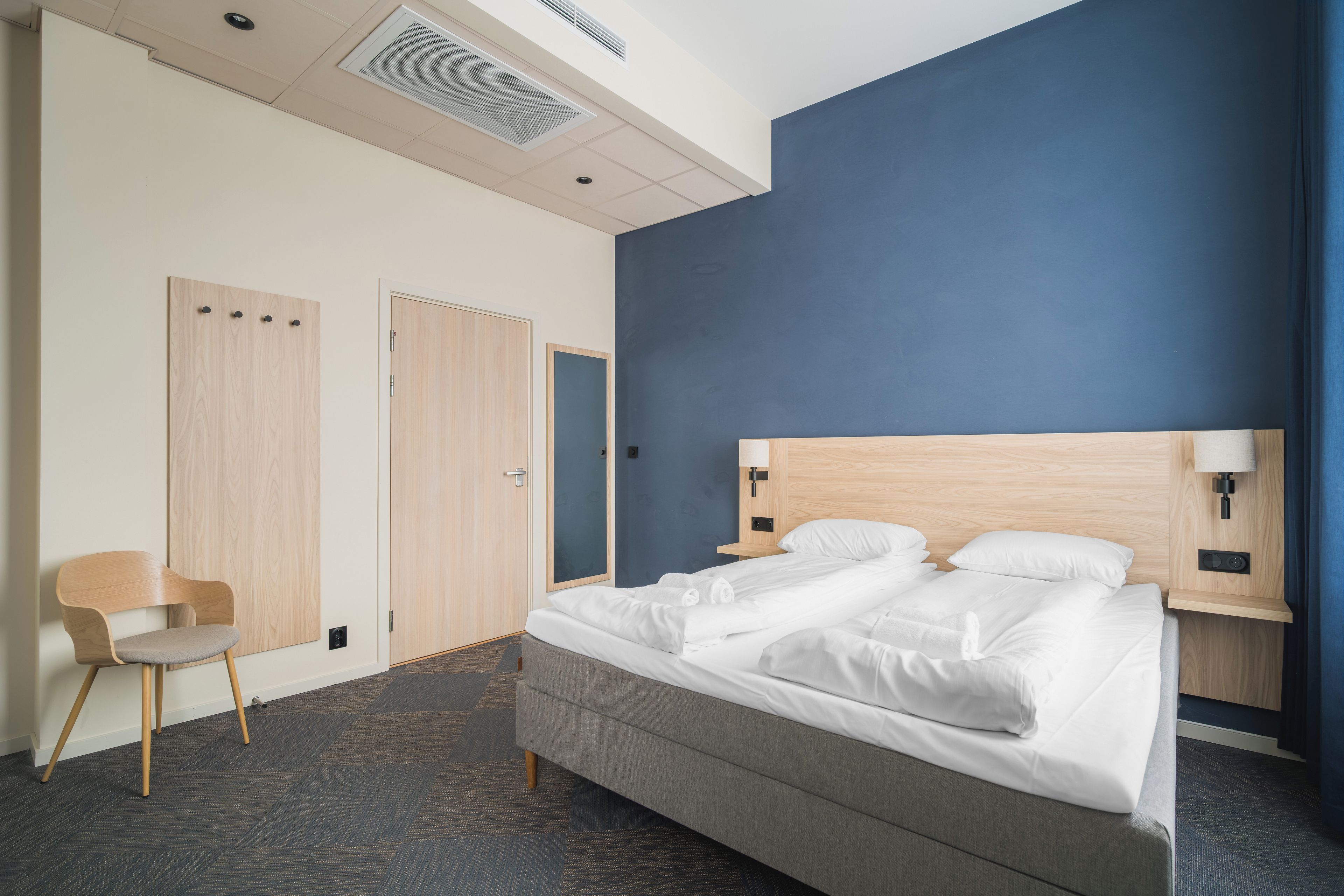 Quarto de hotel com cama, uma cadeira, guarda-roupa e mesa de trabalho com luminária.