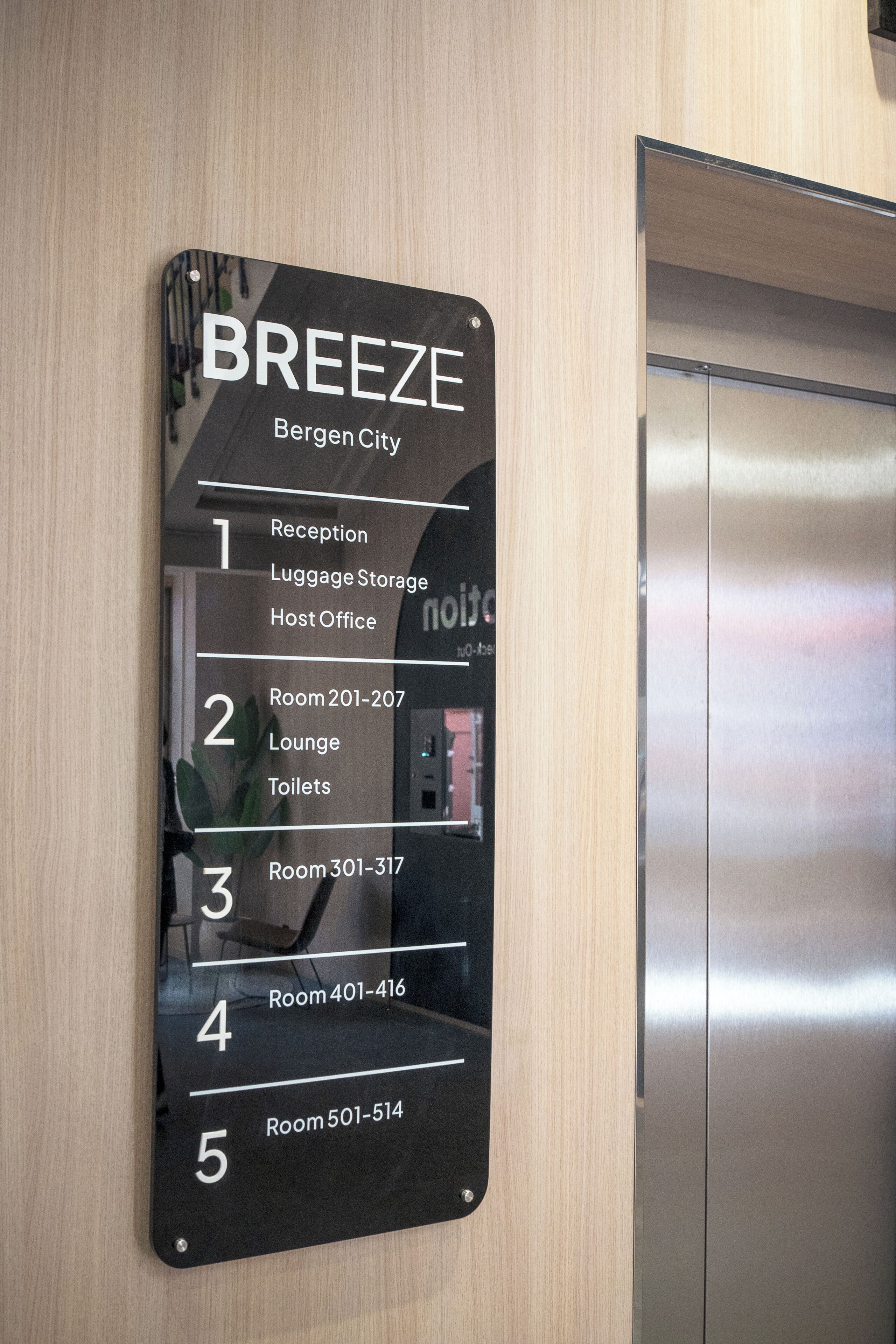 Ein schwarzes Schild mit weißer Schrift, das die Annehmlichkeiten und Zimmernummern des Hotels BREEZE in Bergen City auflistet.