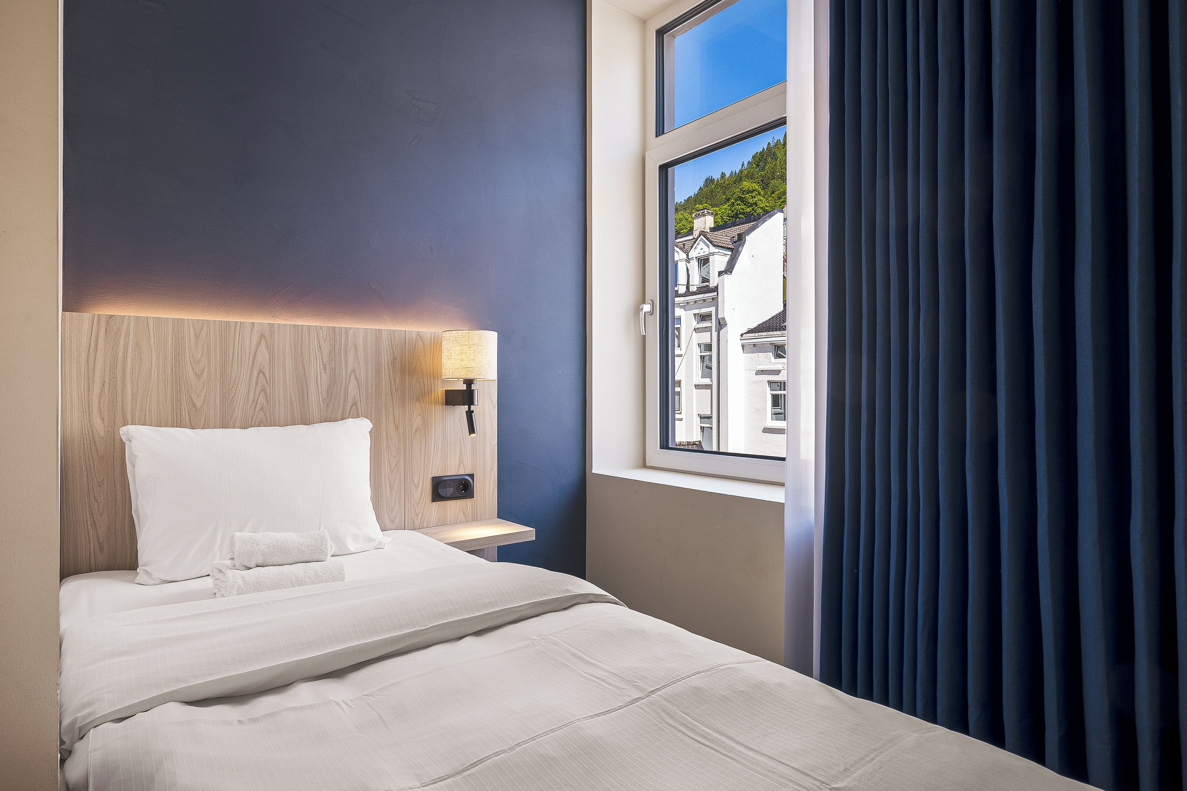 Una camera d'albergo con un letto, un comodino, una lampada e una finestra con tende.