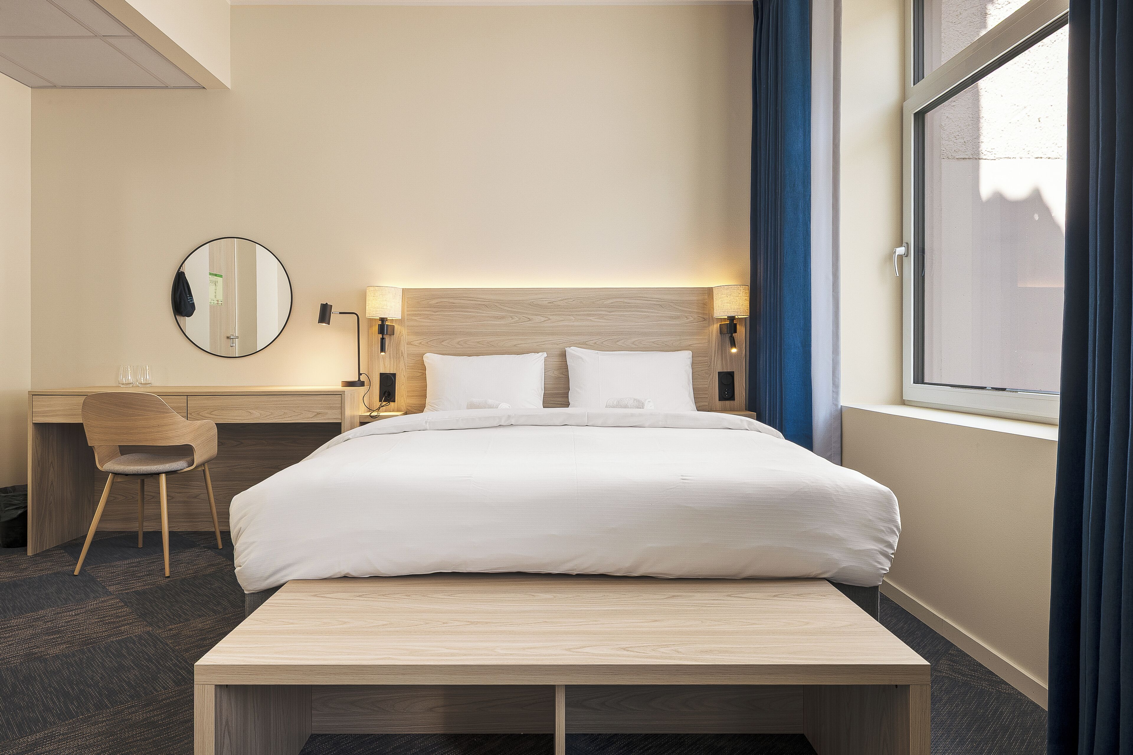 Een hotelkamer met een bed, nachtkastje, lamp, stoel en een groot raam met gordijnen.