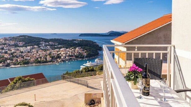 A balcony with a table set for two, overlooking a coastal town and a harbor.