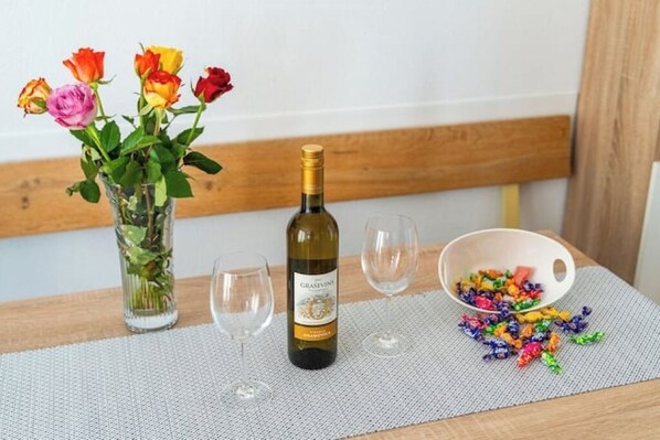 Una botella de vino, dos copas de vino vacías, un cuenco con caramelos de colores, y un jarrón con rosas sobre una mesa.