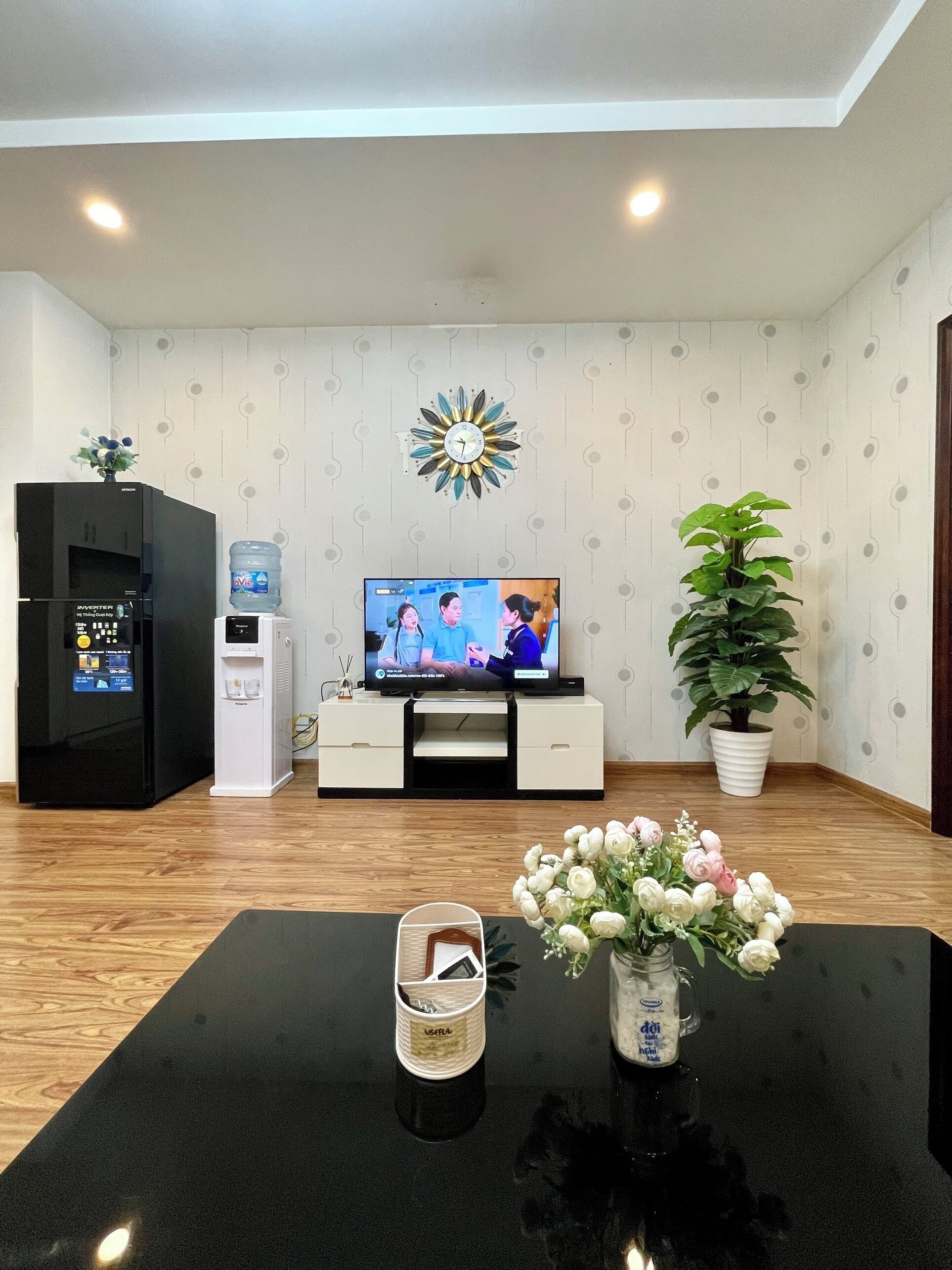 Ein Wohnzimmer mit einem schwarzen Fernseher, einem Wasserspender, einem Kühlschrank, einem weißen Schrank, einer Topfpflanze und einer Vase mit Blumen auf einem schwarzen Tisch.