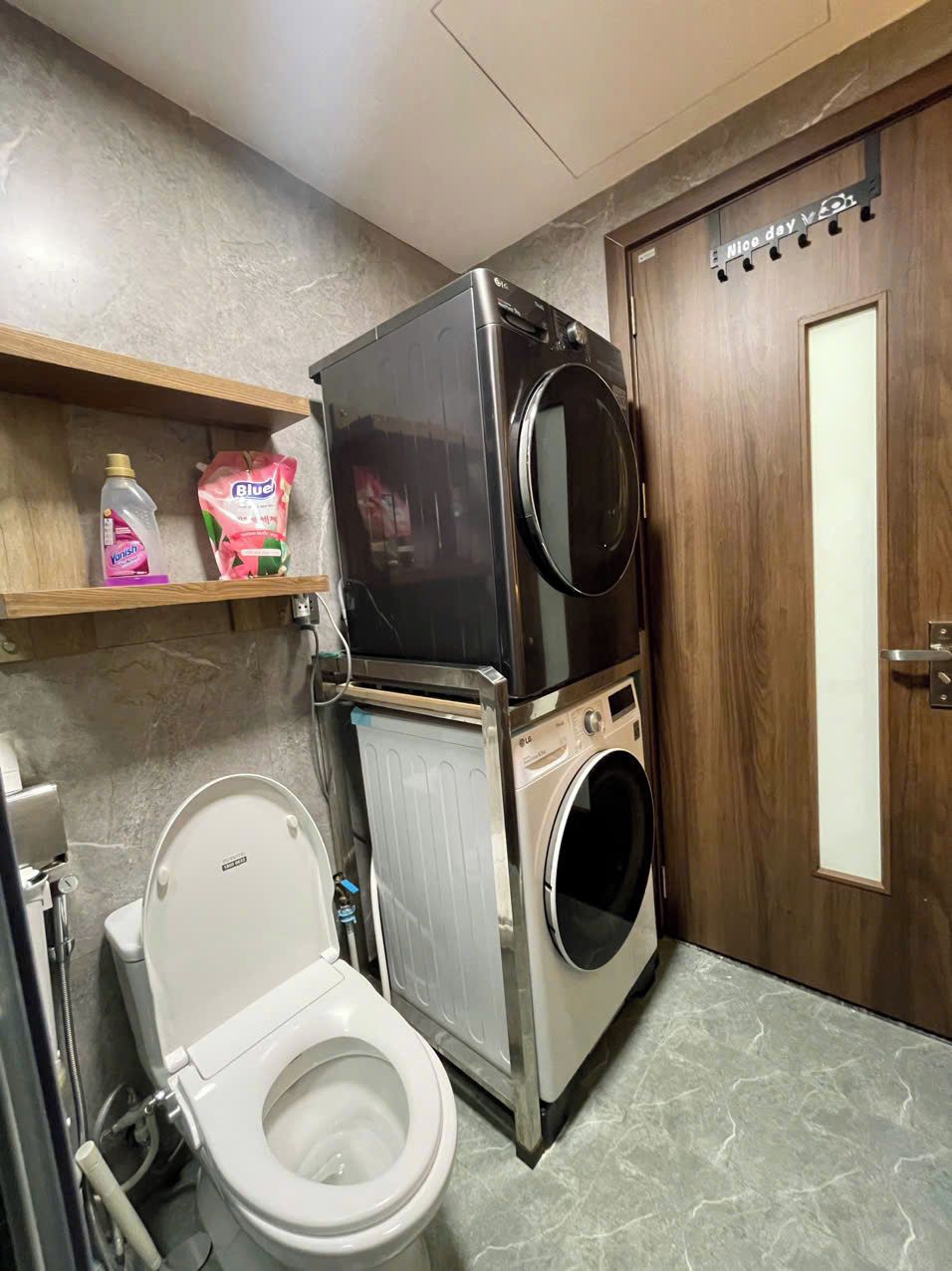 A compact bathroom with a white toilet, a stacked washer and dryer unit, and a shelf with cleaning supplies.