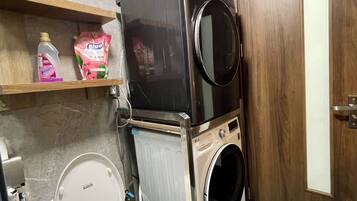 A compact bathroom with a white toilet, a stacked washer and dryer unit, and a shelf with cleaning supplies.