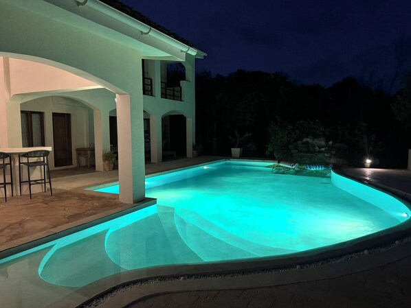 Una piscina con borde curvo, un porche cubierto con sillas y un edificio de dos plantas al fondo.