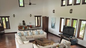A spacious living room with a large white sofa, a wooden coffee table, and a flat-screen TV. There are multiple windows providing natural light and views of greenery outside.