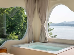 A bathroom with a bathtub, a window with curtains, and a view of a body of water and greenery.