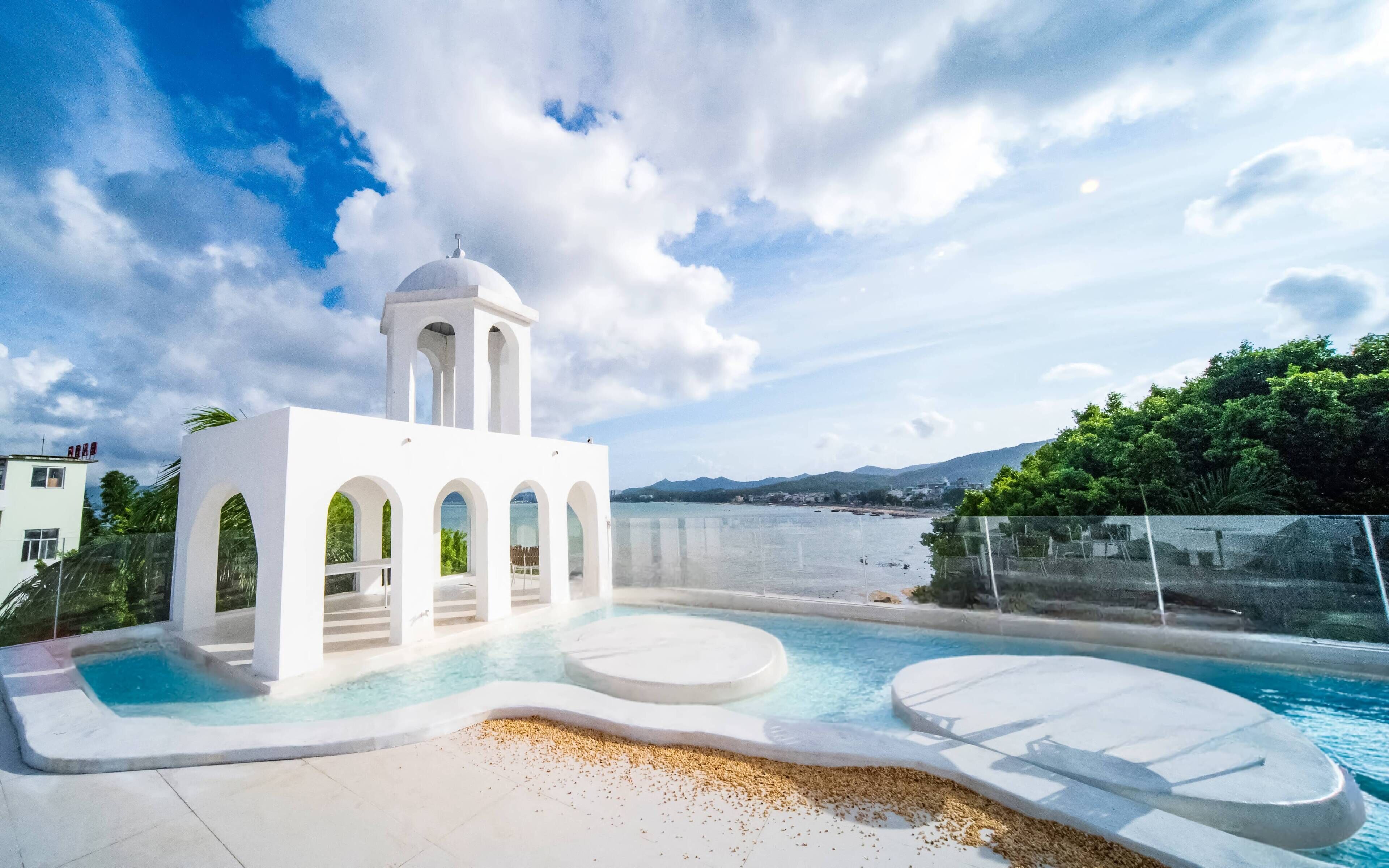 A white building with a bell tower, a pool with a sunken seating area, and a view of the sea and mountains.