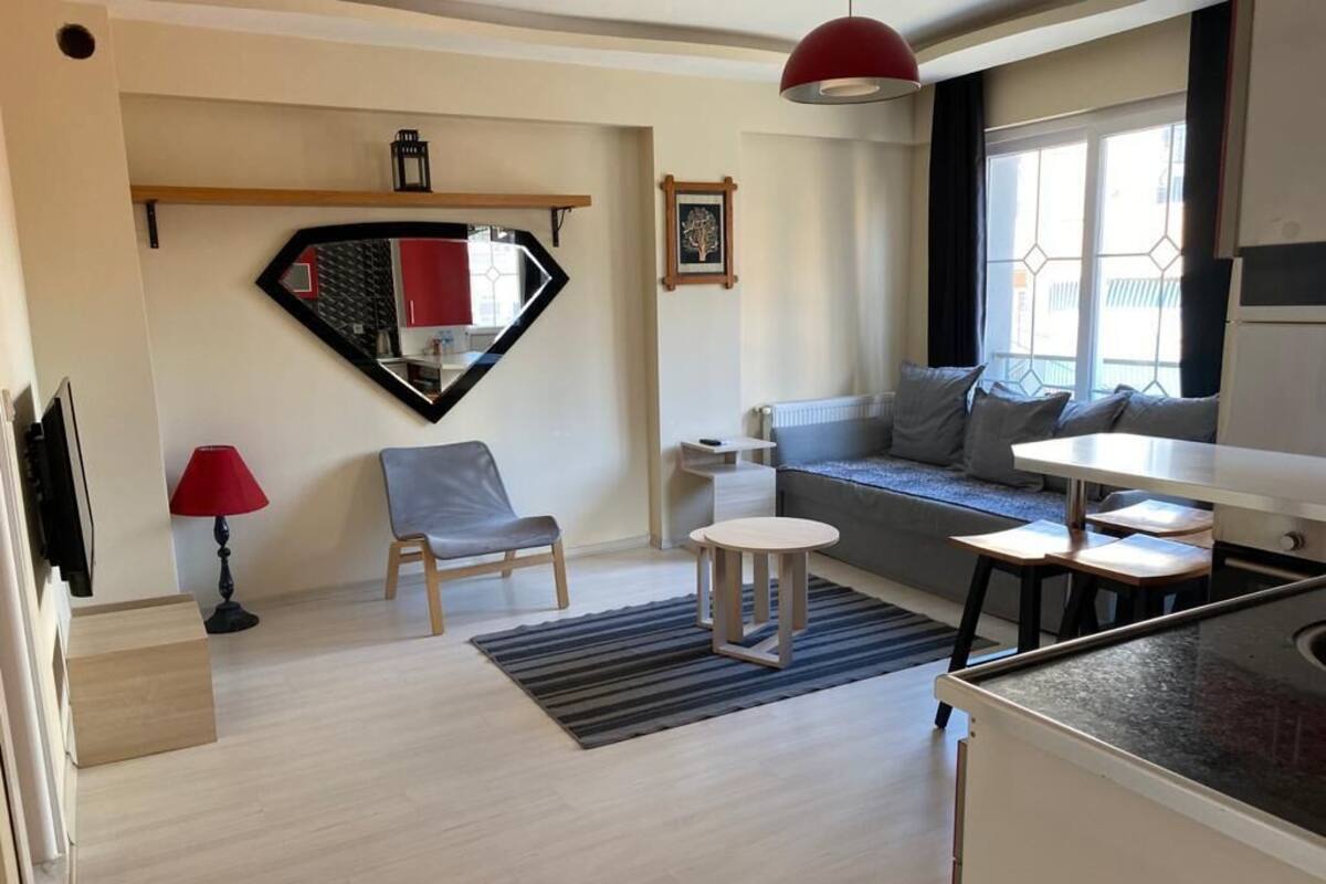 A modern living room with a sofa, a chair, a small table, a red lamp, and a kitchen area with a counter.