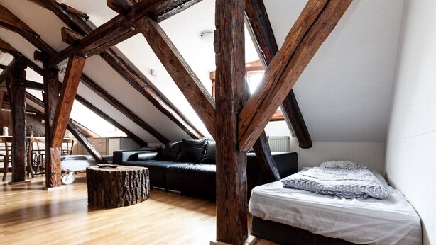A loft bedroom with exposed wooden beams, a wooden floor, a bed, a sofa, and a dining area with a table and chairs.
