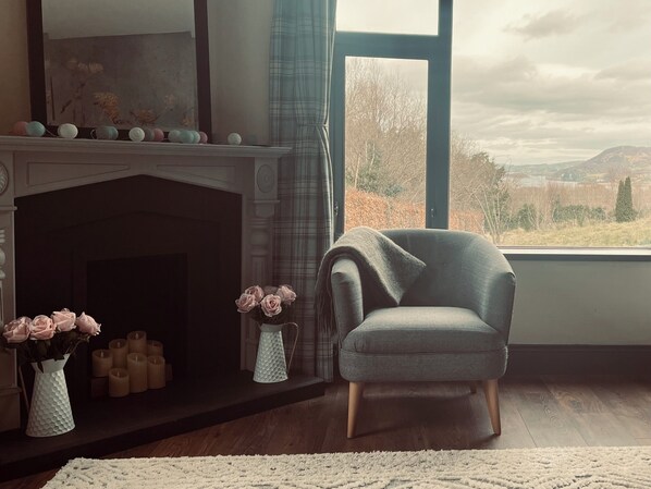 A room with a fireplace, a window with a view of the outdoors, a chair, and a vase with flowers.
