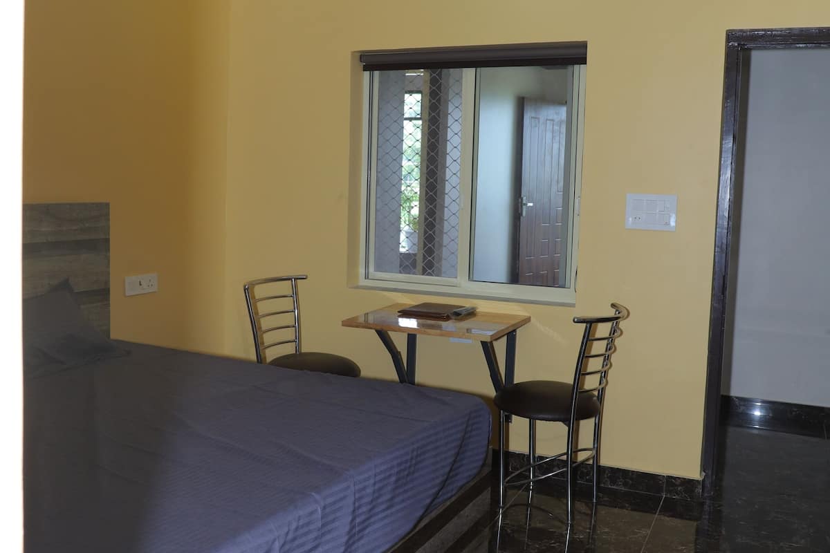 Quarto com cama, mesa, duas cadeiras, ventoinha no teto, janela e porta.