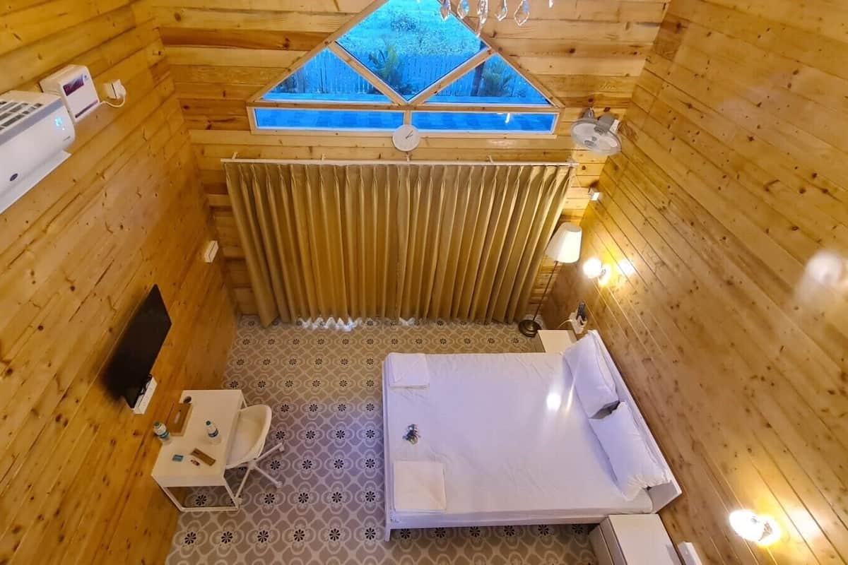 A small, wooden cabin with a bed, a table, a chair, a window, and a wall-mounted heater.