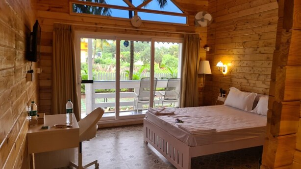 A wooden room with a bed, a desk, and a chair. There is a window with curtains and a view of greenery outside. - Aranya Abode (Arambol)