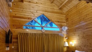 A wooden cabin room with a bed, a desk, a chair, a window with a view, and a chandelier. - Aranya Abode (Arambol)