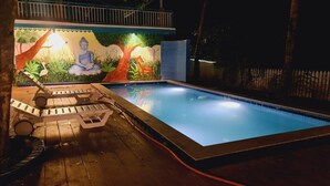 A swimming pool with a mural of a meditating figure and a deer, surrounded by wooden deck chairs and a fence. - Aranya Abode (Arambol)