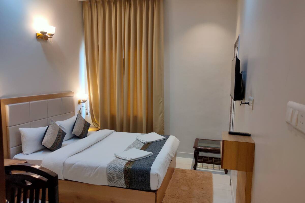 Une chambre d’hôtel avec un lit, une tête de lit en bois, une table de chevet, une télévision et une fenêtre avec des rideaux.