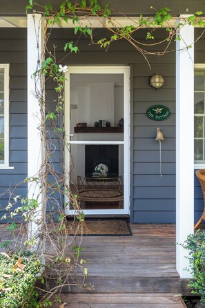 A wooden porch with a white door, a small table with a vase, and a wall-mounted bell. - Ridgedale Cottage, Bayview. The perfect romantic retreat! (Bayview)