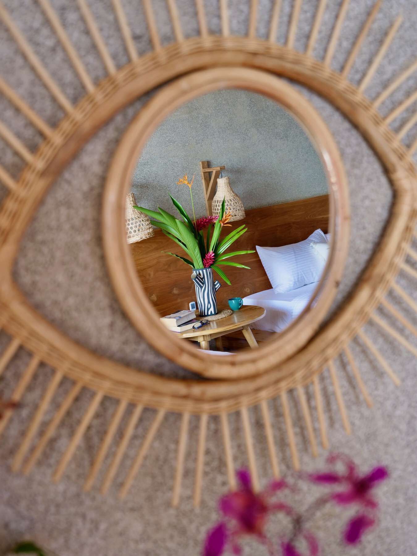 A circular mirror with a sunburst design, reflecting a room with a wooden table, a vase with plants, and a lamp.