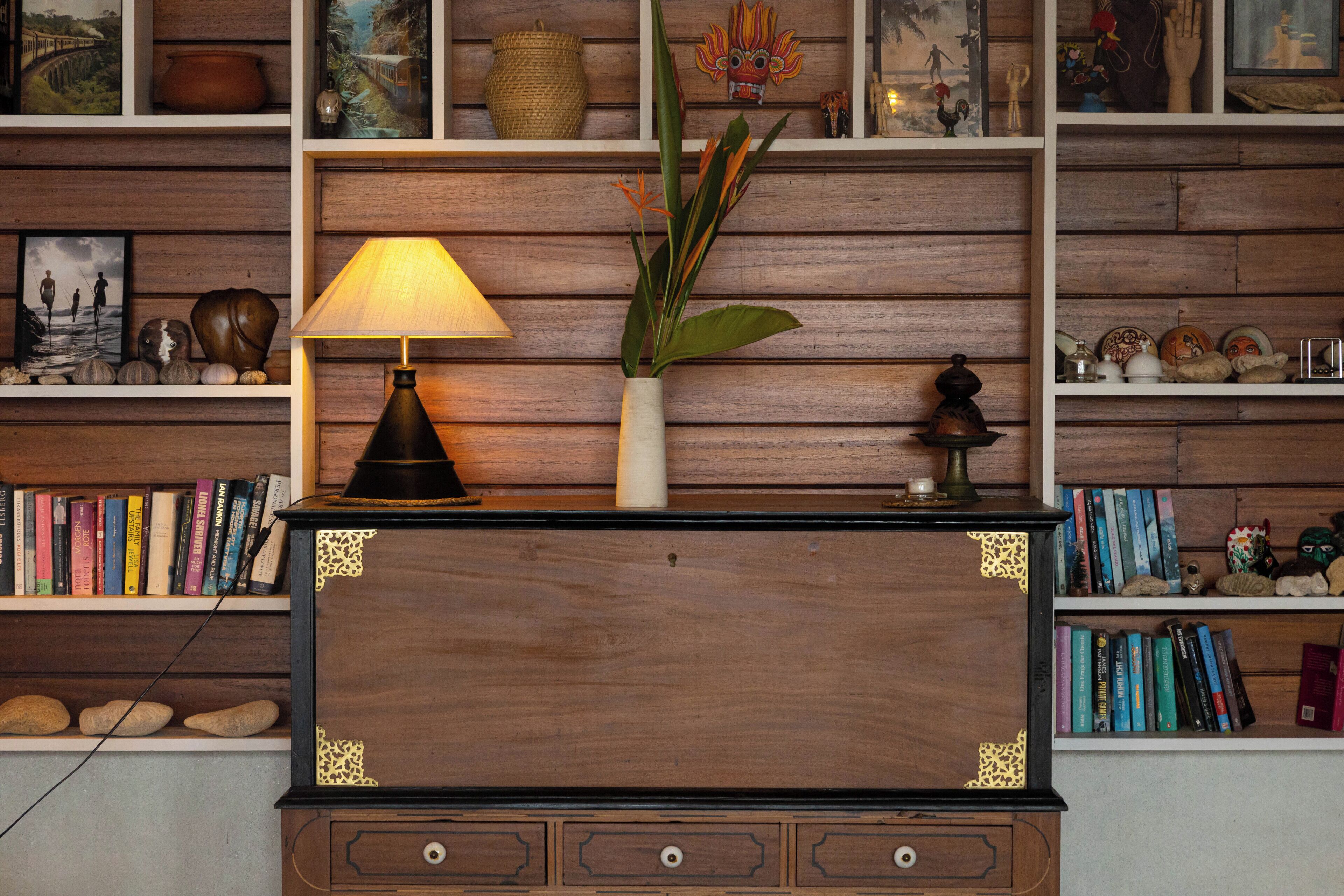 A wooden desk with a lamp, a vase with plants, and shelves with books and decorative items.