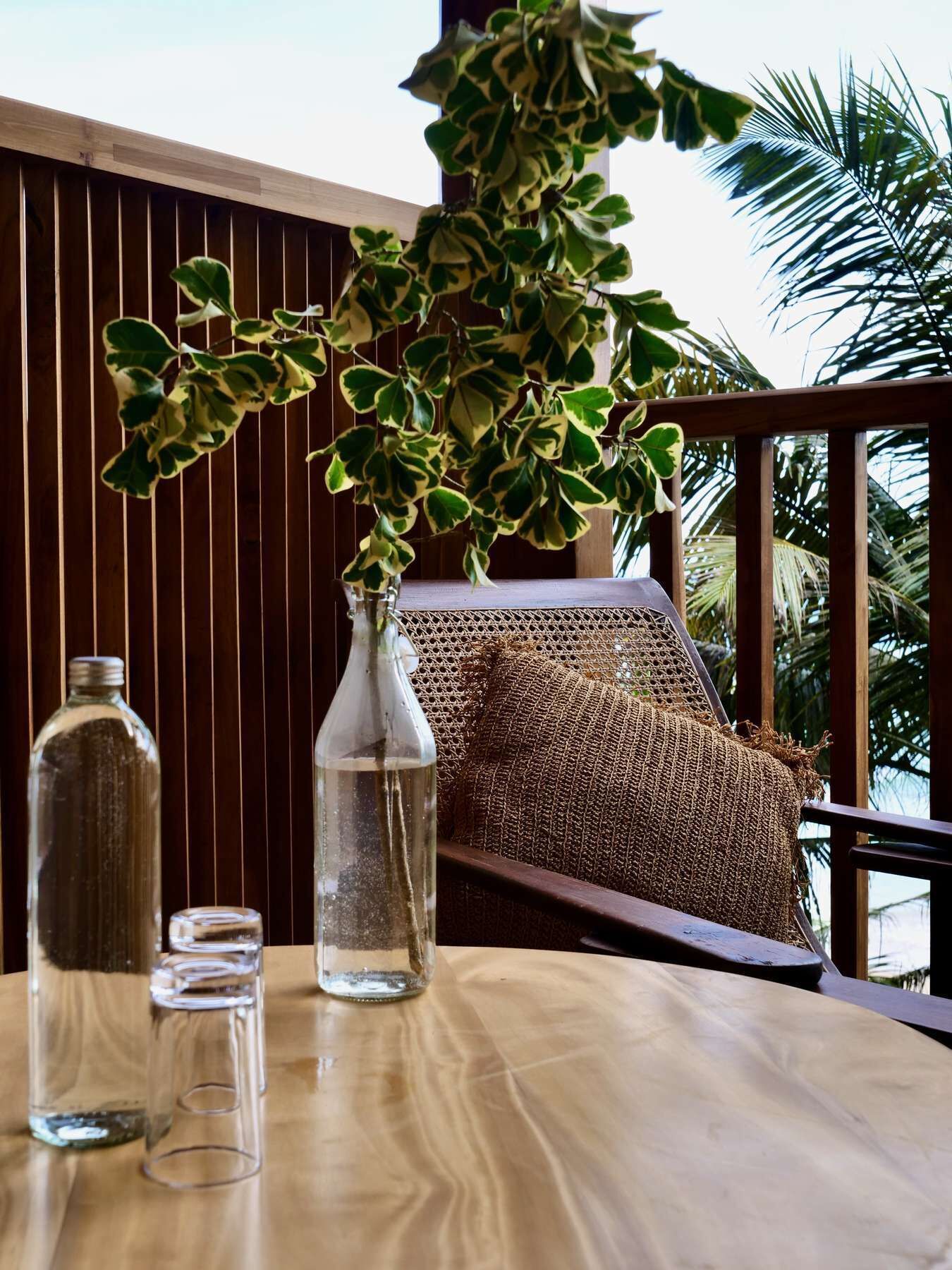 Une table en bois, un vase en verre et des bouteilles, un coussin en fibres tressées, et une vue sur des palmiers.