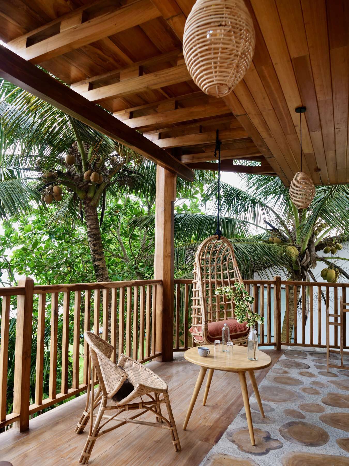 A wooden deck with a wicker chair and a table, surrounded by lush greenery and palm trees.