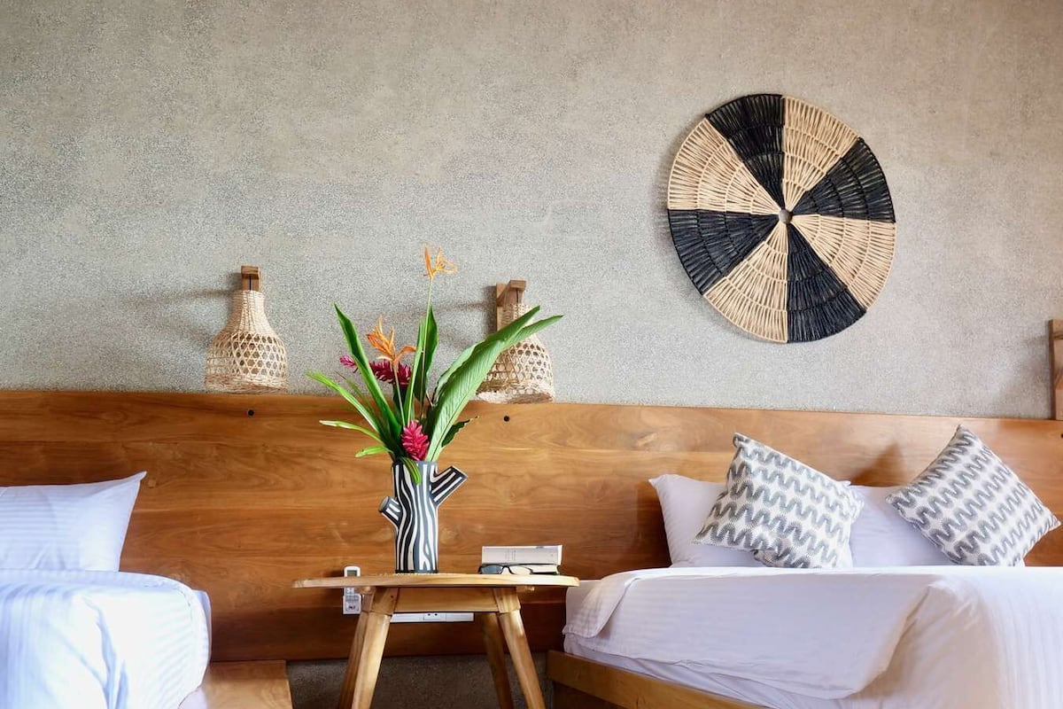 A modern bedroom with a wooden headboard, a round woven wall decoration, a small wooden table with a vase of flowers, and a blue throw blanket.
