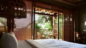 A bedroom with a bed, a chair, and a table. There is a window with blinds and a view of palm trees outside. - Coast 67 Boutique Hotel (Ahangama)