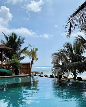 Zona de piscina con vistas al océano, rodeada de palmeras y una terraza de madera.