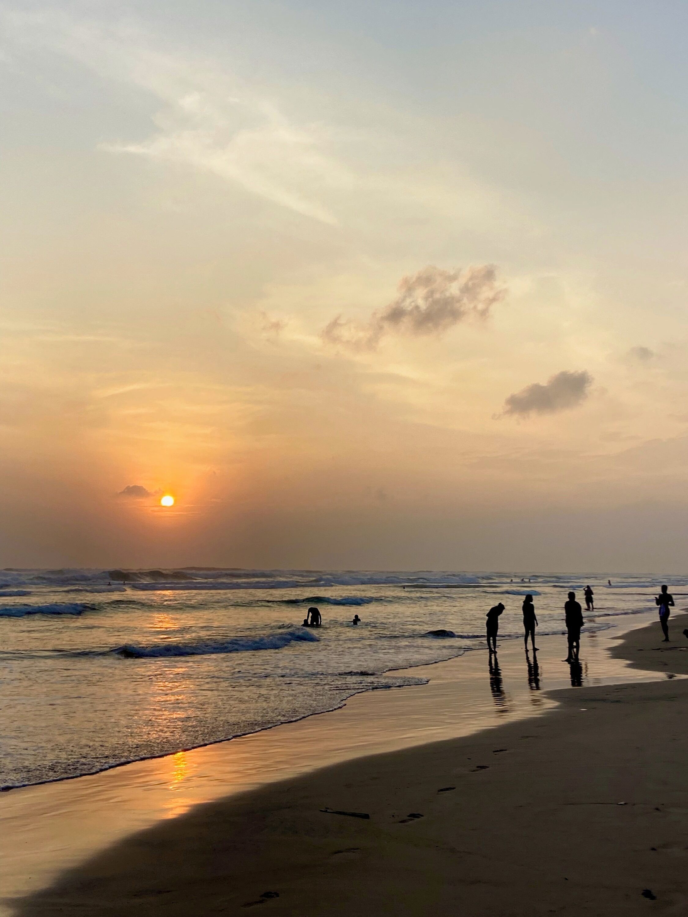 Escena de playa con el sol poniéndose sobre el océano, gente caminando por la orilla y olas rompiendo en la playa.