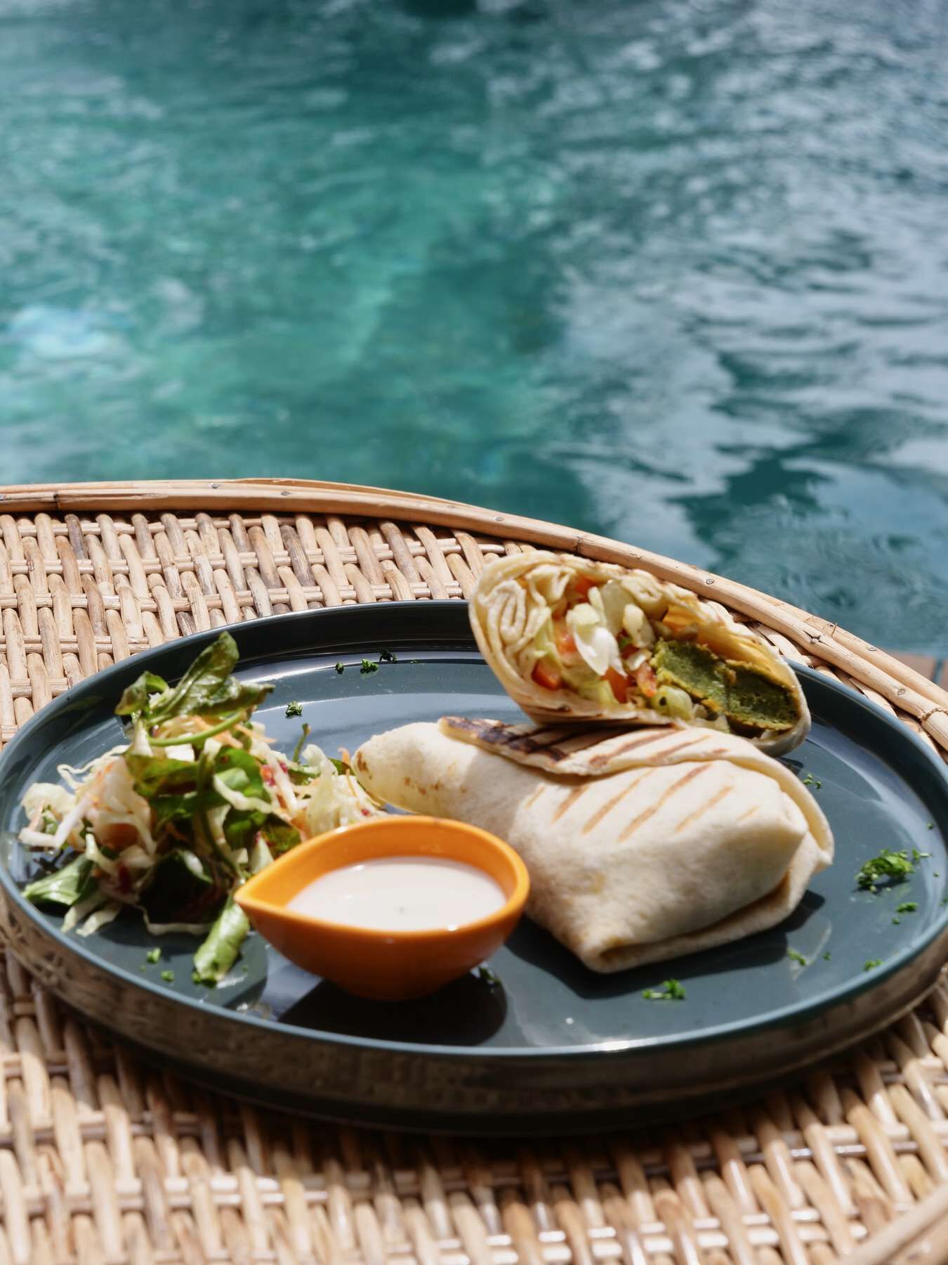 Un plato con un wrap, una ensalada y un recipiente pequeño de salsa sobre una mesa de mimbre junto a una piscina.