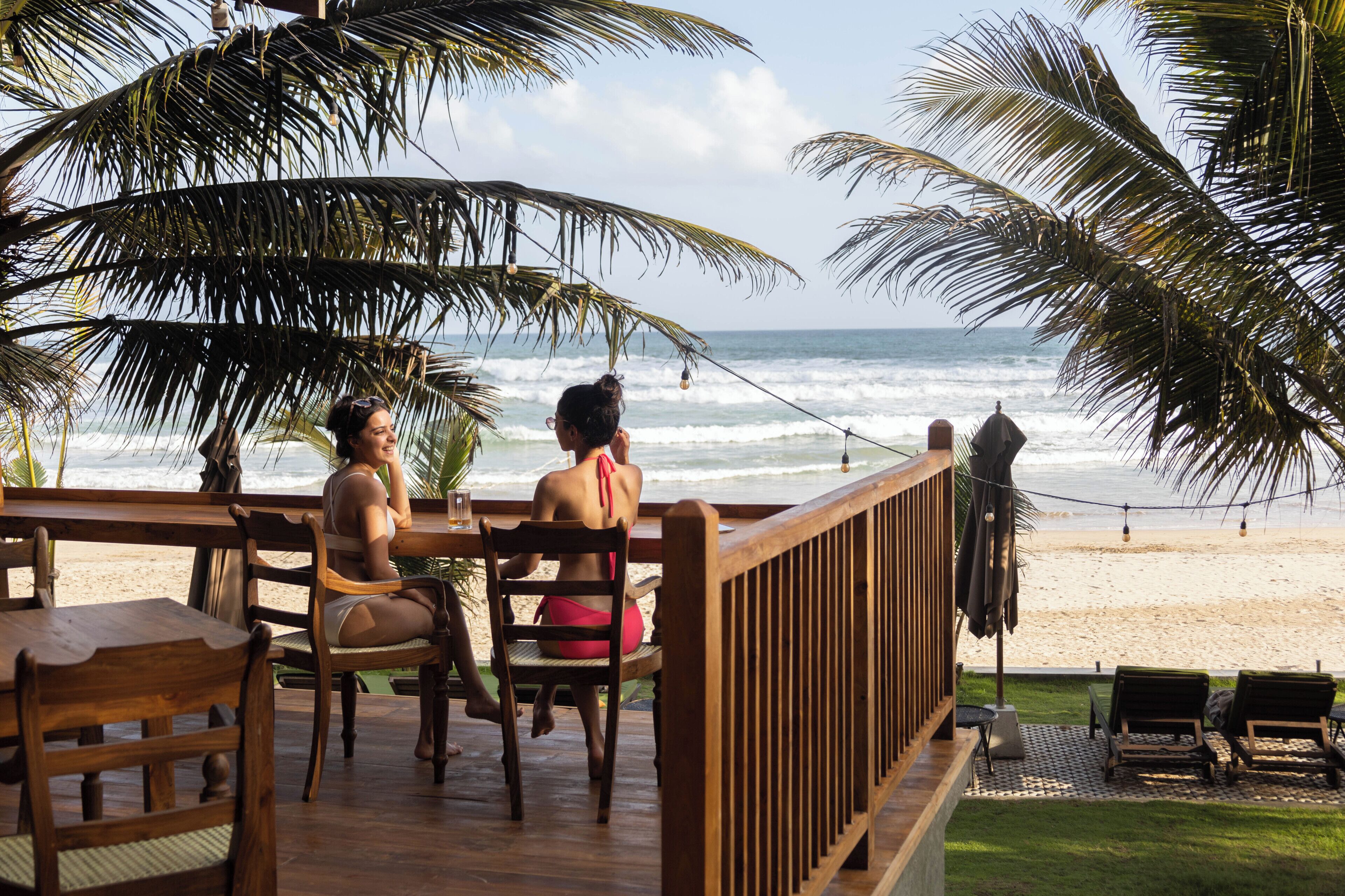 A wooden deck with chairs overlooking a beach, palm trees, and a view of the ocean.