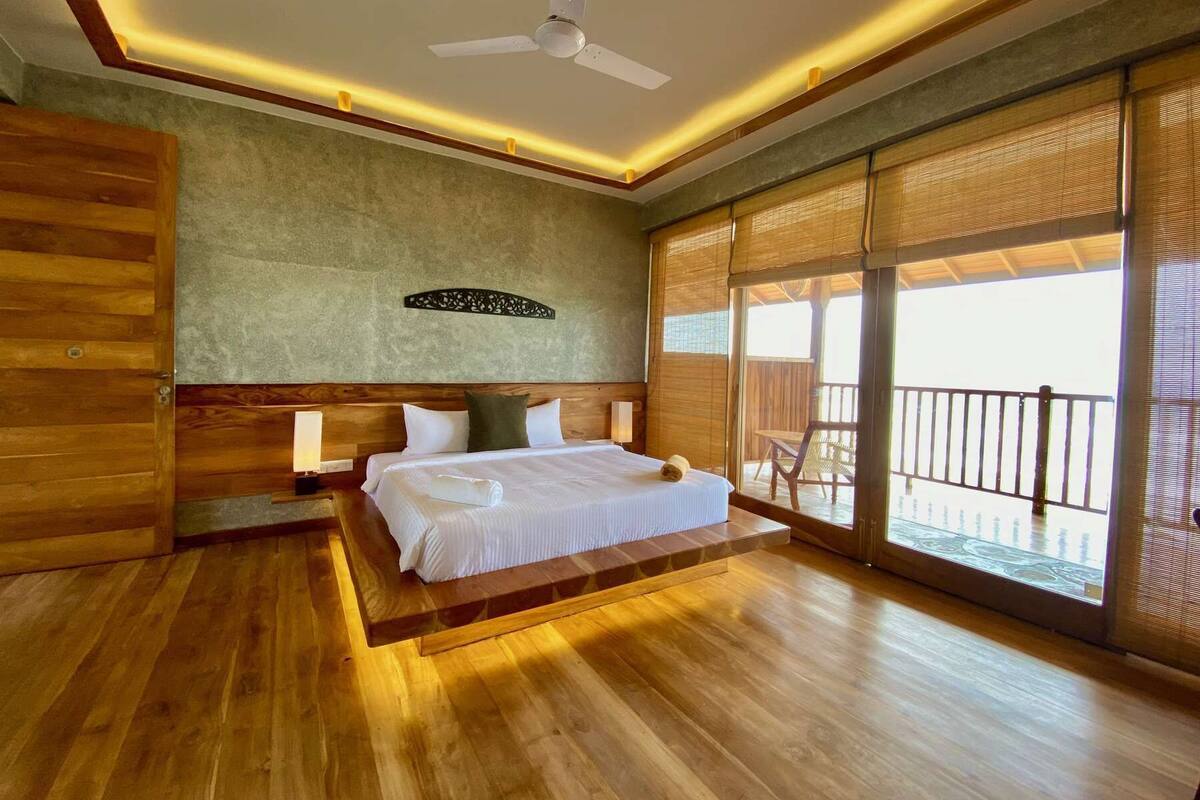 A hotel room with a wooden floor, a bed with white linens, a ceiling fan, and a large sliding glass door leading to an outdoor balcony.
