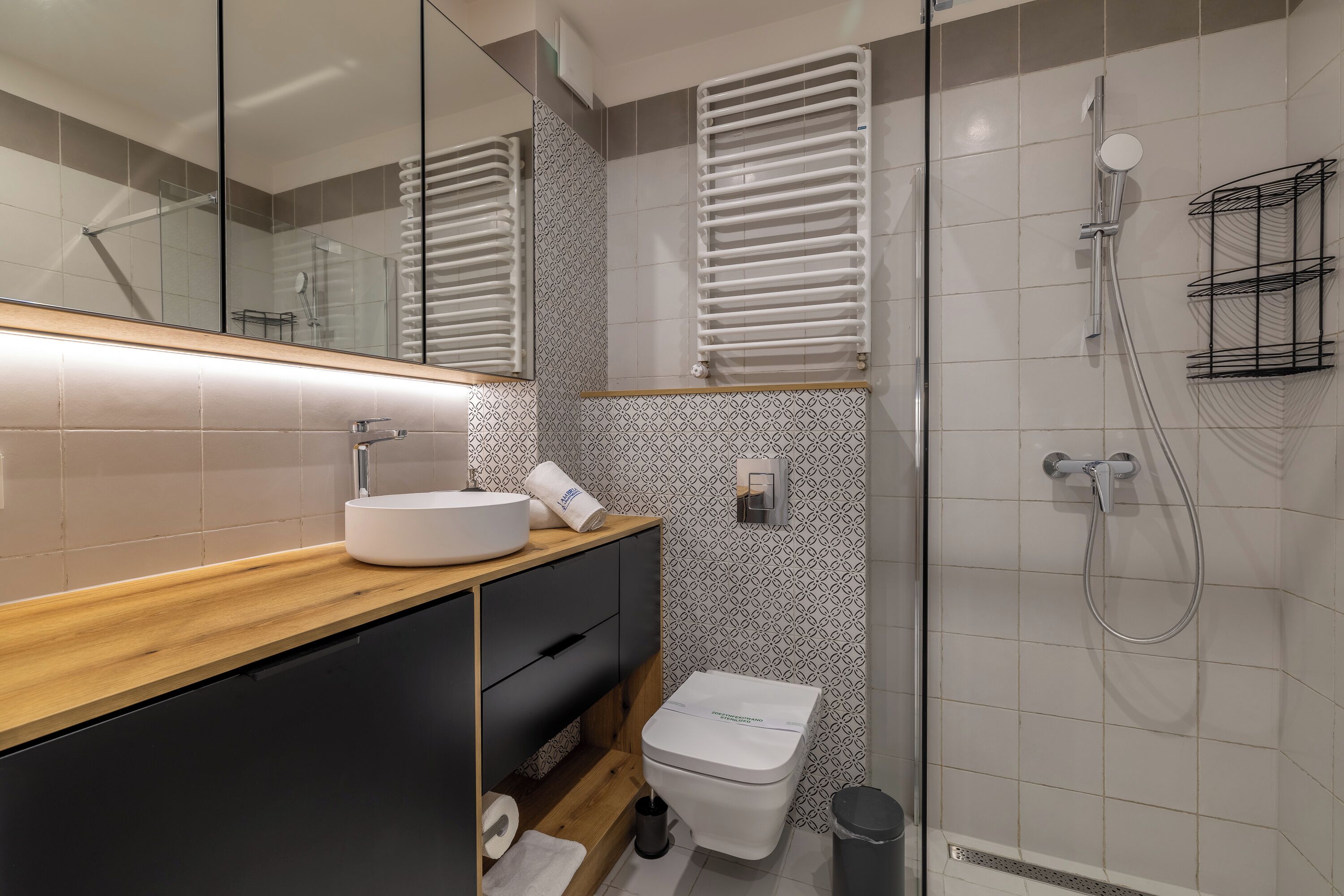 Un baño moderno con tocador de madera, un lavabo blanco, inodoro y zona de ducha con mampara de vidrio.