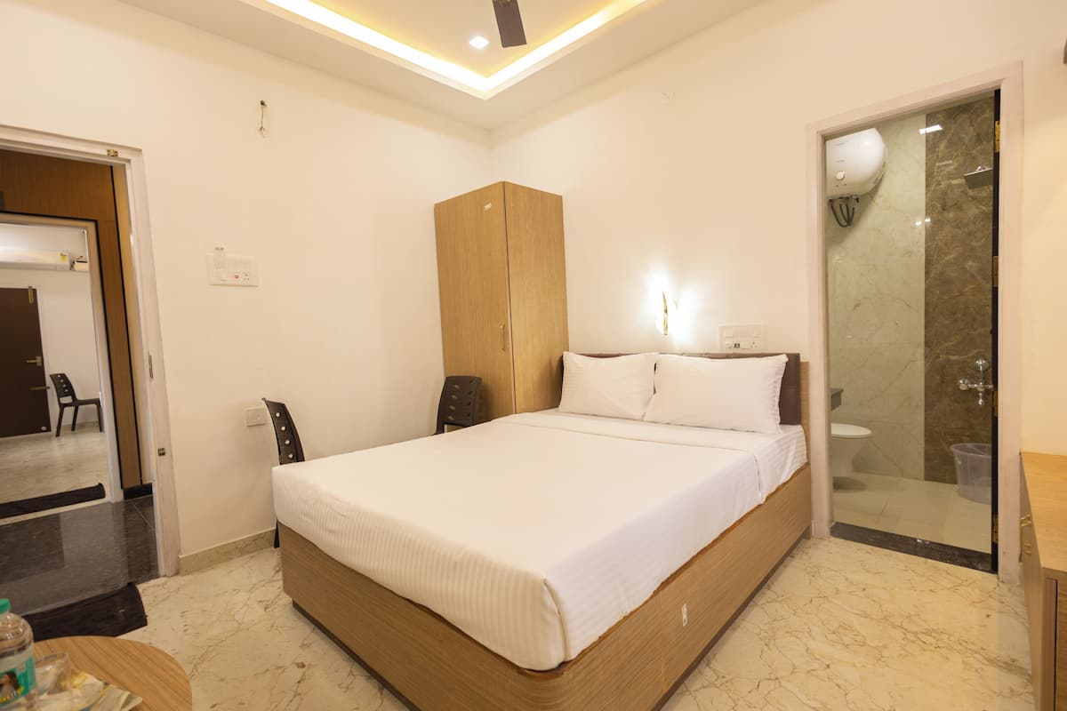 A neatly arranged bedroom with a large bed, a wooden wardrobe, a glass shower enclosure, and a small table with a chair.
