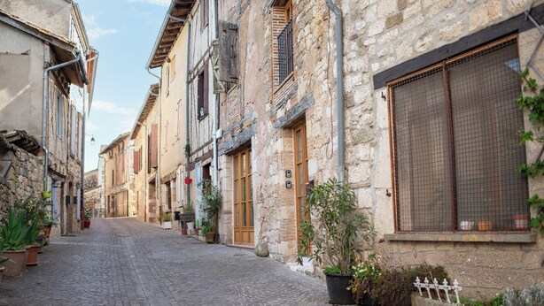 Une rue pavée bordée de bâtiments historiques en pierre, de plantes en pot et d’une ruelle étroite.
