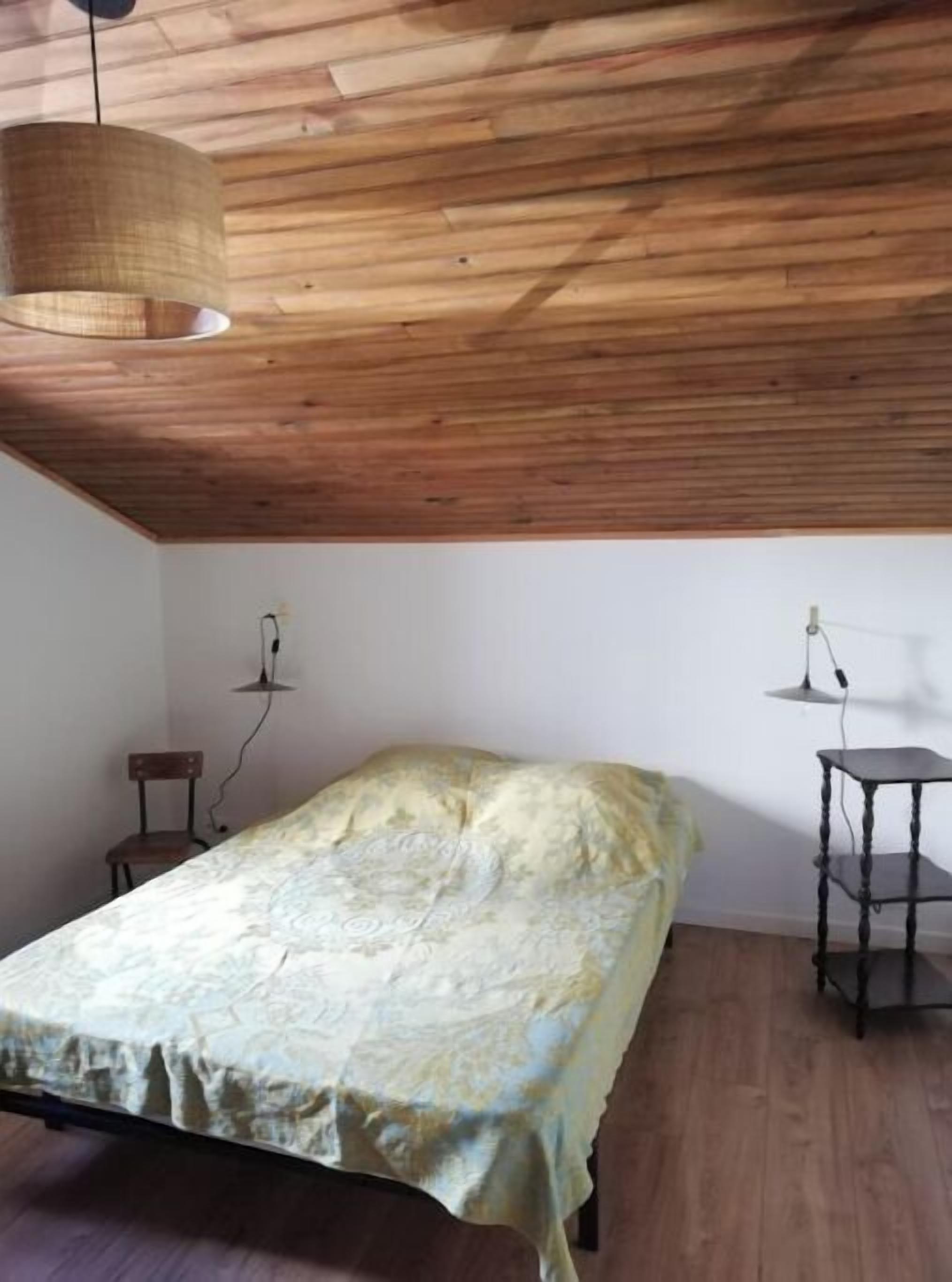 Un dormitorio con techo de madera, una cama con colcha estampada, una pequeña silla de madera y una mesita de noche con lámpara.