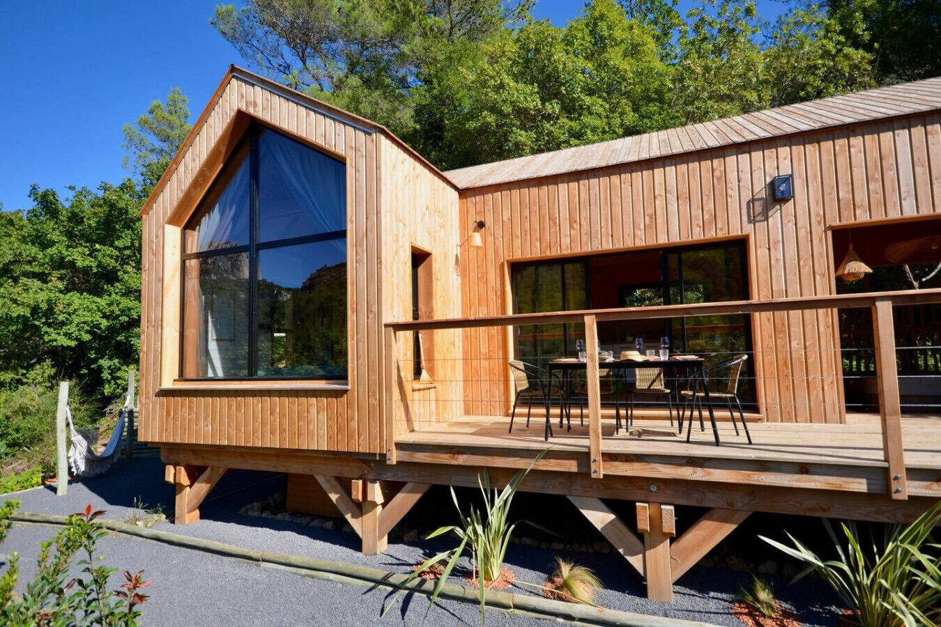 Une cabane en bois dotée de grandes fenêtres, d’une terrasse avec des meubles d’extérieur et entourée d’un cadre naturel.