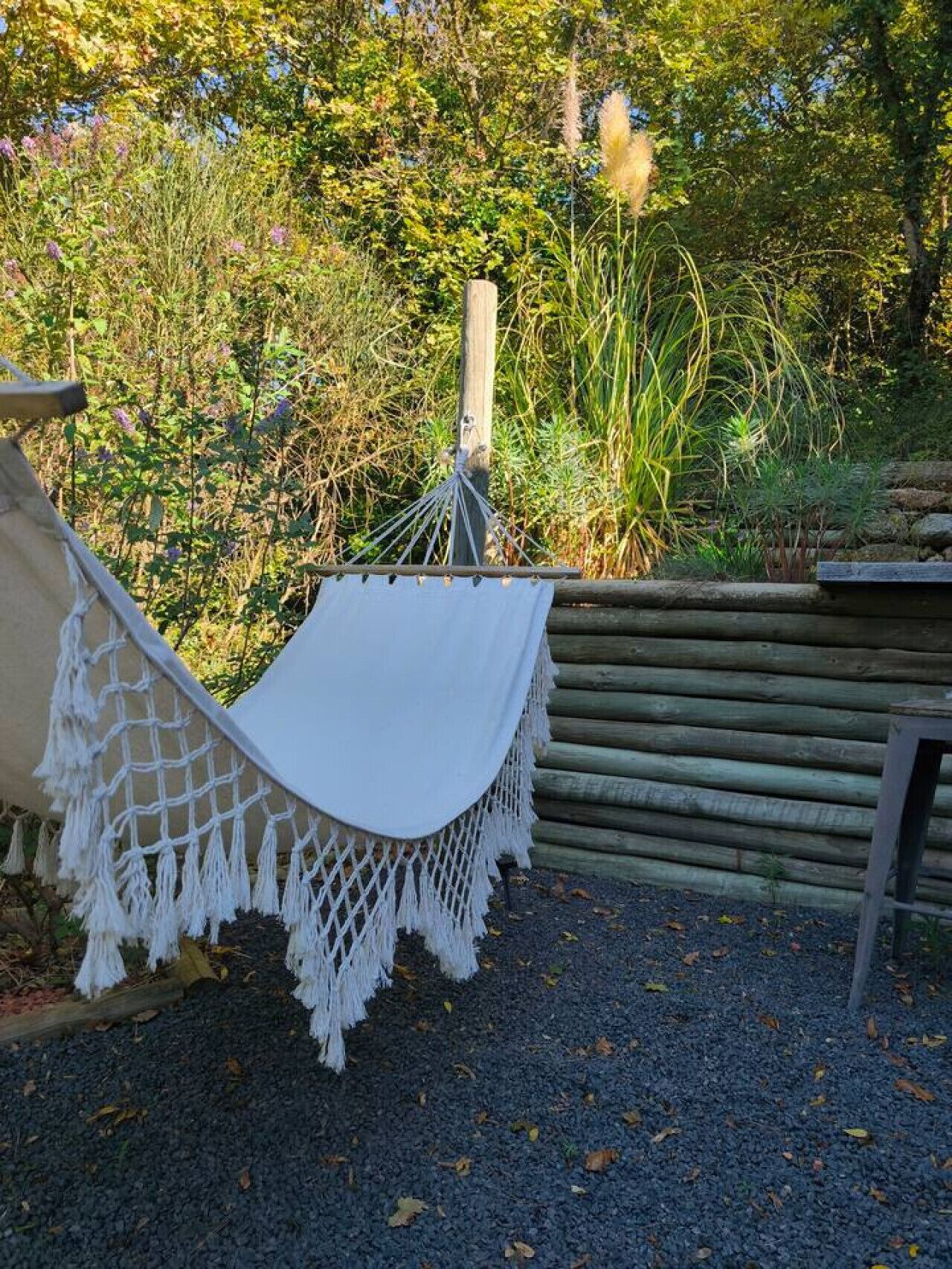 Un hamac est attaché à un poteau en bois dans un jardin, lequel est agrémenté d’un chemin de gravier et d’une clôture en bois.