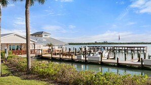 A marina with docked boats, a building with a covered patio, and a grassy area with shrubs. - The Riverside Club - B204 (Marco Island)