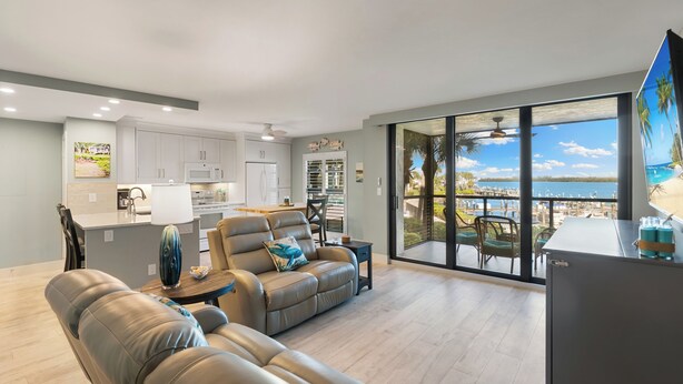 A modern living room with a sofa and a dining area, a kitchen with white cabinets, and a large sliding glass door leading to a balcony with a view of the ocean. - The Riverside Club - B204 (Marco Island)